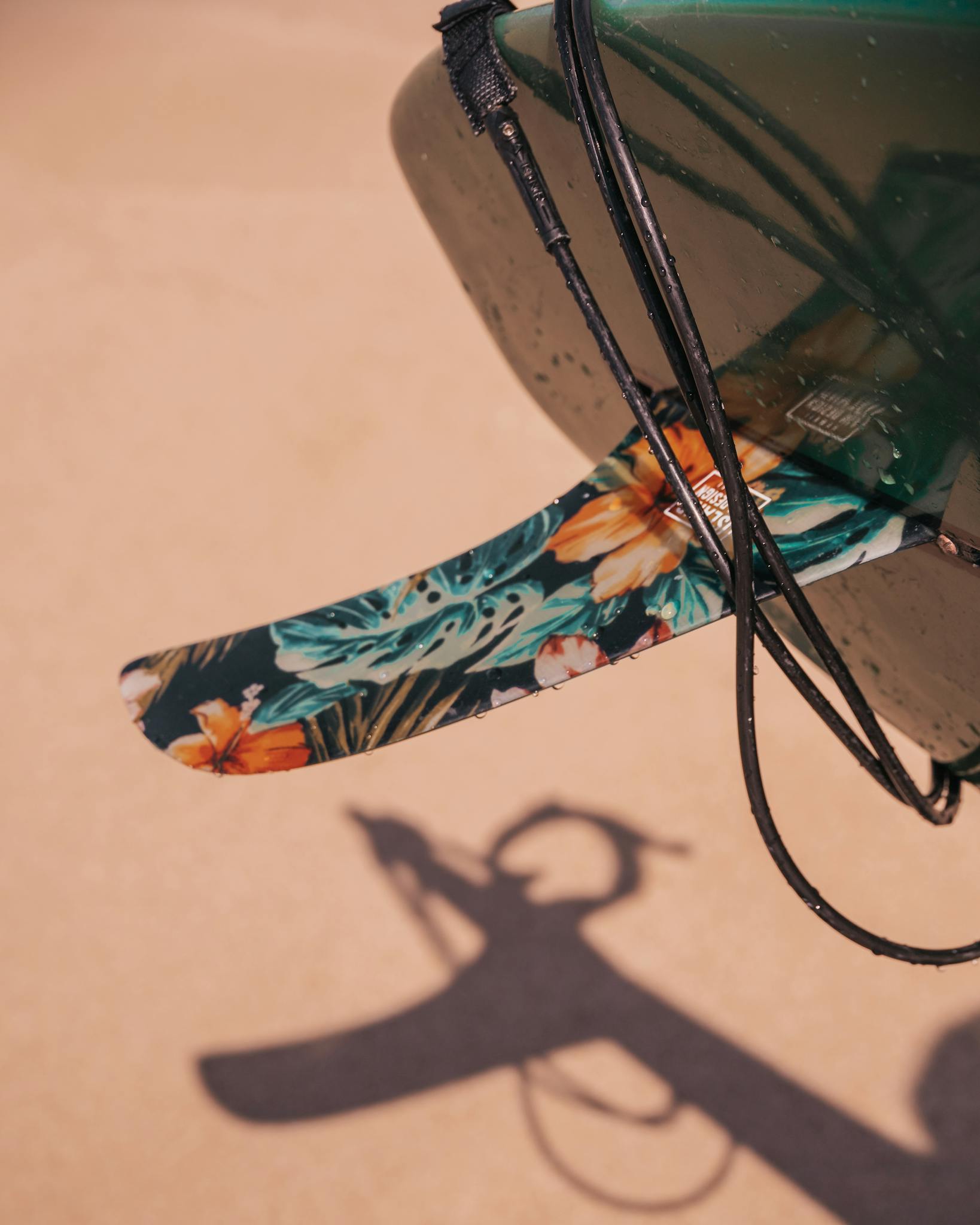 Close-up of a floral surfboard fin casting a shadow on a sandy Hawaiian beach.